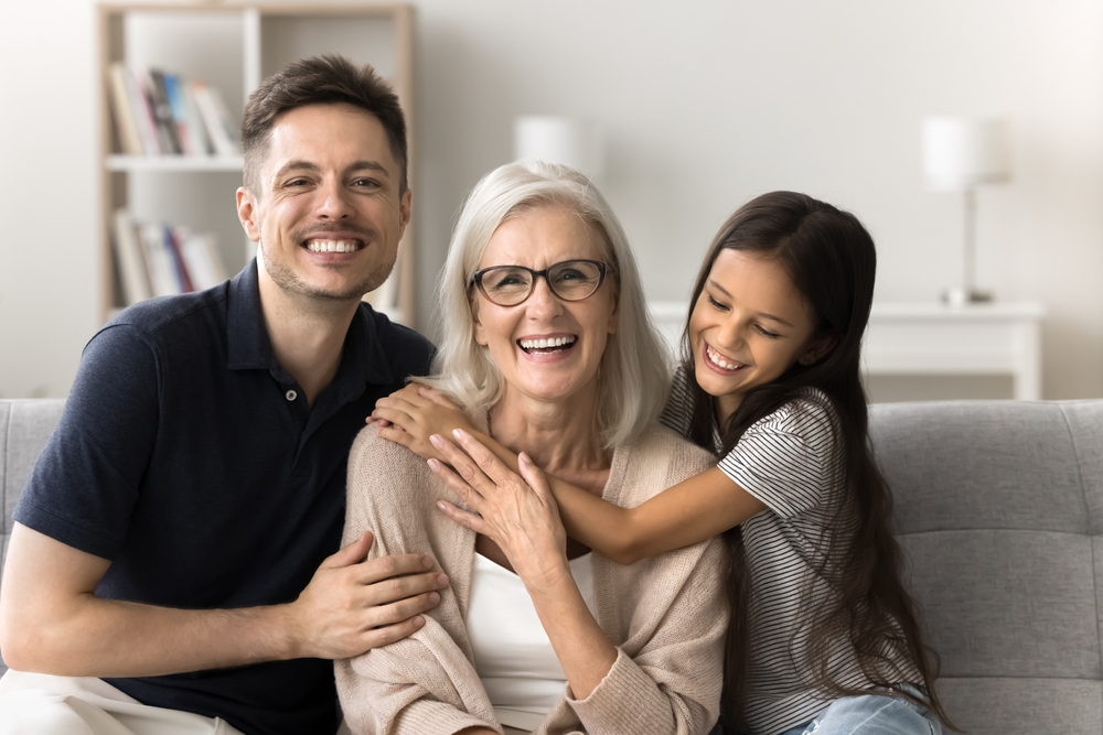 A smiling man, a senior woman with glasses, and a young girl sitting together on a couch, highlighting healthy teeth and family bonding in a bright living room.
